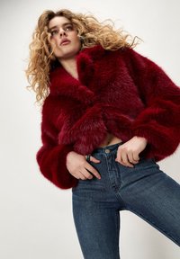 Mujer joven con cabello rizado que lleva una chaqueta de piel sintética rojo profundo y pantalones vaqueros azul oscuro de tiro alto, posando con las manos cerca de la cintura.