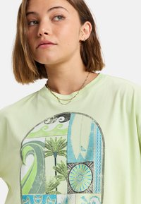 Camiseta de color verde claro con un diseño gráfico multirrizado que presenta olas, palmeras y motivos de tablas de surf en acentos azules y negros.
