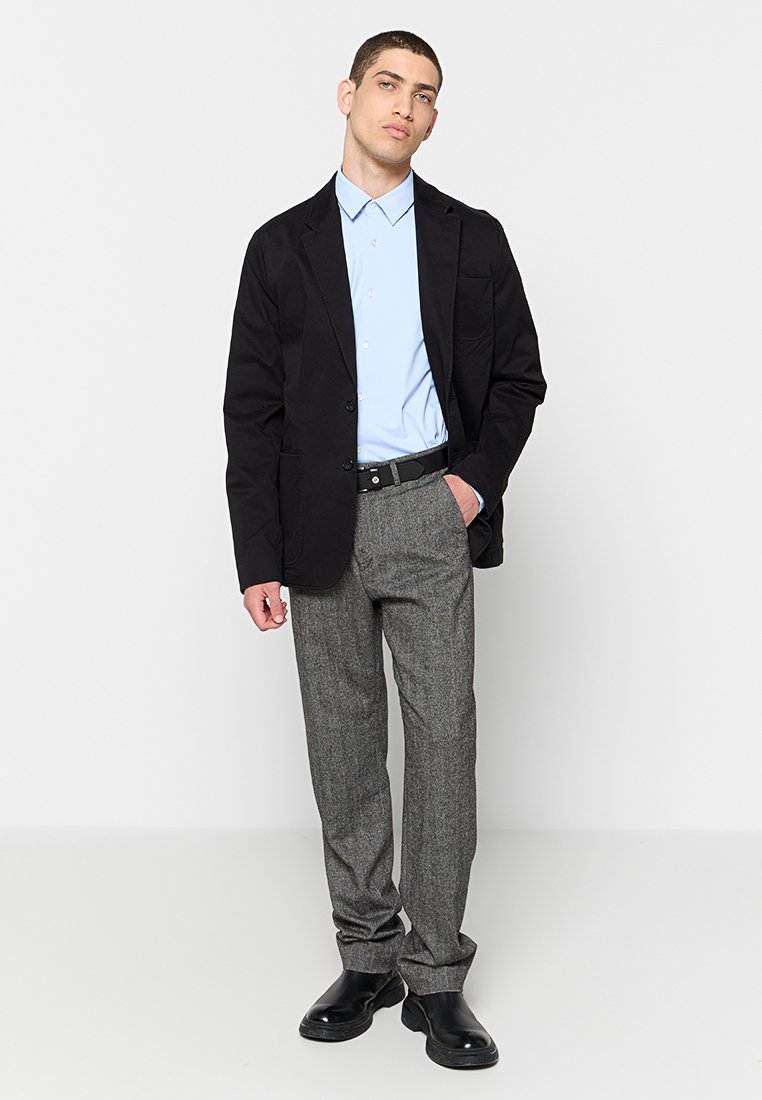 Homme portant un blazer noir, une chemise bleu clair, un pantalon gris, une ceinture noire et des bottes noires, se tenant avec une main dans la poche.