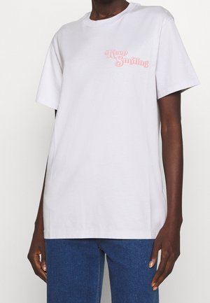 Henkilö, jolla on valkoinen t-paita, jossa on vaaleanpunaisella tekstillä "Keep Smiling" rinnassa, ja siniset farkut, seisoo yksinkertaisen taustan edessä.