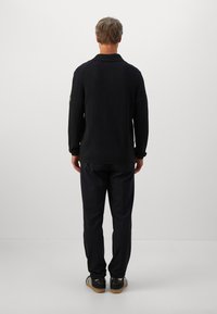 Hombre de pie frente a una pared blanca, vistiendo un suéter negro de canalé, pantalones oscuros y zapatillas negras con rayas blancas y suelas de goma.
