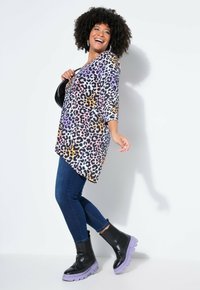 Frau mit lockigen Haaren, die lächelt, trägt ein buntes Oberteil mit Leopardenmuster, dunkle enge Jeans, schwarze Stiefel mit lila Sohlen und trägt eine schwarze Handtasche.