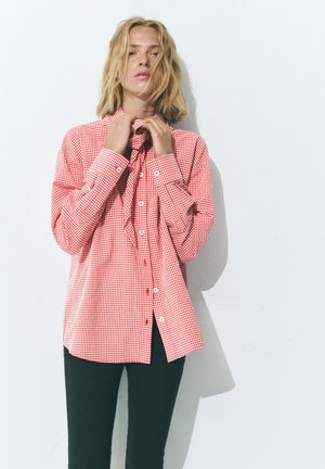 Blonde Frau passt ein rot-weiß kariertes Hemd mit Knöpfen an, trägt schwarze Hose und steht an einer weißen Wand.