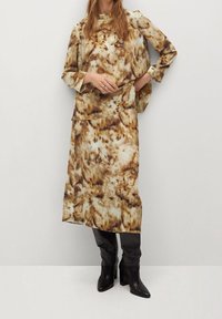 Braun und beige gemustertes Kleid mit langen Ärmeln und hohem Kragen, schichtweise gestaltet. Getragen mit schwarzen kniehohen Stiefeletten mit Absatz.
