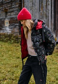 Prešívaná čierna péřová bunda s kvetinovým vzorom, červenou podšívkou a kapucňou. Béžový sveter s vrkočovým vzorom a čierne nohavice. Červená pletená čiapka.