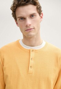 Suéter de punto amarillo con cuello redondo, que presenta una tapeta de botones con tres botones. Textura suave, diseño casual.