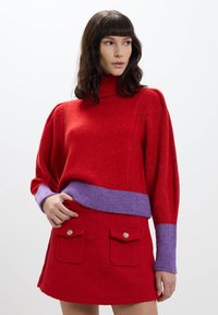 Maglione rosso lavorato a maglia con un alto colletto e una banda viola in vita, abbinato a una gonna rossa con due pattine per le tasche e bottoni decorativi.