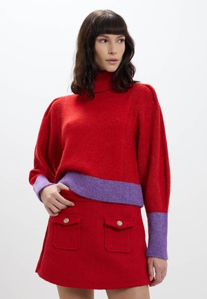 Maglione rosso lavorato a maglia con un alto colletto e una banda viola in vita, abbinato a una gonna rossa con due pattine per le tasche e bottoni decorativi.