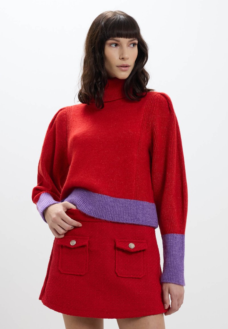 Maglione rosso lavorato a maglia con un alto colletto e una banda viola in vita, abbinato a una gonna rossa con due pattine per le tasche e bottoni decorativi.