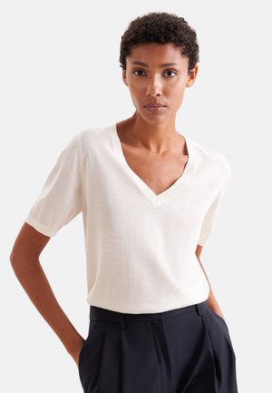 Persona con capelli corti e ricci che indossa una maglietta bianca a maniche corte con scollo a V e pantaloni neri con le mani nelle tasche.
