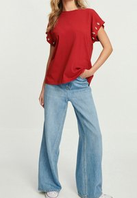 Vrouw in een rood t-shirt met korte mouwen, versierd met pareltjes op de mouwen, en een lichtblauwe broek met wijdere pijpen, gecombineerd met witte sneakers.