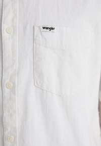 Camicia bianca in lino con chiusura frontale a bottoni, taschino a sinistra con etichetta "Wrangler" e bottoni bianchi. Tessuto liscio, design informale.