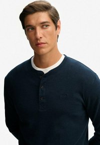 Jonge man met kort bruin haar, gekleed in een marineblauwe henley shirt over een wit onderhemd, die naar links kijkt tegen een effen witte achtergrond.