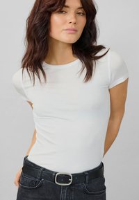 Maglietta bianca aderente con maniche corte, realizzata in tessuto morbido. Indossata con jeans scuri a vita alta e una cintura nera con una fibbia argentata.