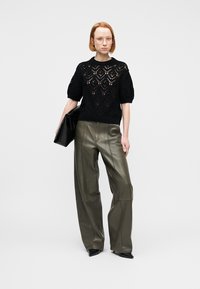 Femeie purtând un pulover croșetat negru cu mâneci scurte, pantaloni largi din piele de culoare măslinie, pantofi negri cu vârf ascuțit, ținând o poșetă plic neagră.