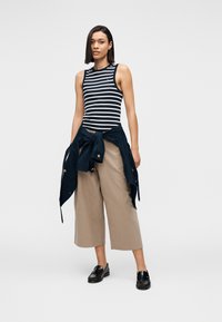 Ριγέ αμάνικη μπλούζα σε ναυτικό μπλε και λευκό, συνδυασμένη με μπεζ παντελόνι φαρδιάς γραμμής και δεμένο ναυτικό μπλε σακάκι. Μαύρα μοκασίνια ολοκληρώνουν το σύνολο.