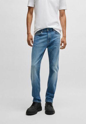 Mann trägt hellblaue gerade Jeans, schwarze Schnürstiefel und ein weißes Kurzarm-T-Shirt vor einem schlichten Hintergrund.