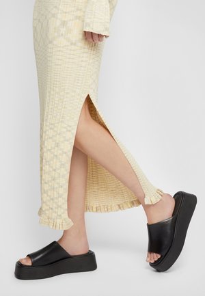 Sandali piattaforma neri con punta aperta, abbinati a un vestito giallo a trama con uno spacco laterale e dettagli di frange all'orlo.