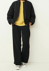 Kurtka bomber z pikowanej czarnej tkaniny na żółtym swetrze dzianinowym, w połączeniu z czarnymi spodniami o szerokich nogawkach i szarymi sneakersami na białej podeszwie.