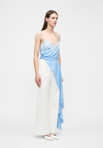 Γυναίκα που στέκεται φορώντας μπλε ασύμμετρο τοπ με δαντέλα, διάκοσμο με ανάγλυφη λεπτομέρεια, λευκό παντελόνι με φαρδιά πόδια και λευκές γόβες.