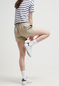 Beige šortky s elastickým pasem, spojené s bílým tričkem se navy proužky a bílými teniskami s černými akcenty, umístěné na neutrálním pozadí.