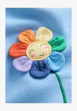 Hymyilevä kangaskukka monivärisillä terälehdillä vaaleansinisellä kankaalla, jossa oranssin, keltaisen, vihreän, sinisen ja violetin sävyjä.