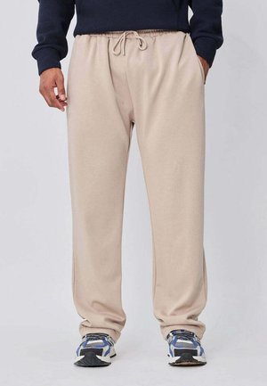 Pantalon de survêtement beige à cordon porté avec des chaussures de course bleues, grises et noires, les mains posées négligemment dans les poches, sur un fond uni.