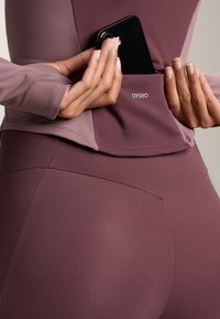 Burgundi sportfelső hosszú ujjakkal és testhezálló leggingssel, zsebből telefon számára. Az anyag simának és rugalmasnak tűnik.