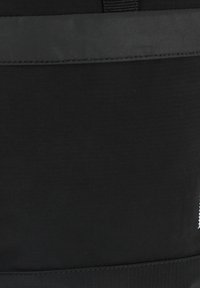 Bolsa de tela negra con una textura suave, que presenta un diseño de rayas horizontales y una cremallera en la parte superior. El logo de la marca es parcialmente visible.