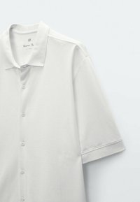 Weißes Polohemd mit kurzen Ärmeln aus Baumwolle. Verfügt über eine Knopfleiste und einen klassischen Kragen. Weiche Textur mit minimalistischem Design.