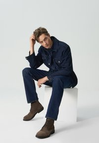 Ciemnoniebieska dżinsowa kurtka o wyraźnej strukturze, dopasowane dżinsy i brązowe zamszowe buty na sznurówki. Model siedzi na białym tle w kształcie sześcianu.