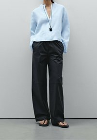 Henkilö, joka käyttää löysää vaaleansinistä paitaa, mustia leveälahkeisia housuja, kädet taskuissa, mustaa riipuskorua ja mustia flip-flop-sandaaleja.