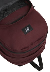 Mochila color burdeos hecha de tejido texturizado, con cierre superior de cremallera, parche de marca y un bolsillo delantero con cremallera.
