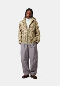 Giacca con motivo camouflage in beige, caratterizzata da una chiusura con zip, cappuccio regolabile e multiple tasche frontali. Indossata con pantaloni cargo grigi larghi.