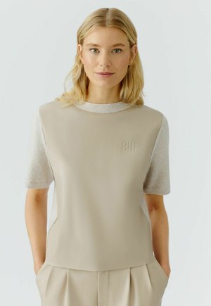 Beige Oberteil mit kurzen Ärmeln, das ein glattes Vorderteil und strukturierte pulloverähnliche Ärmel aufweist. Dezentes Logo-Detail auf der Brust.