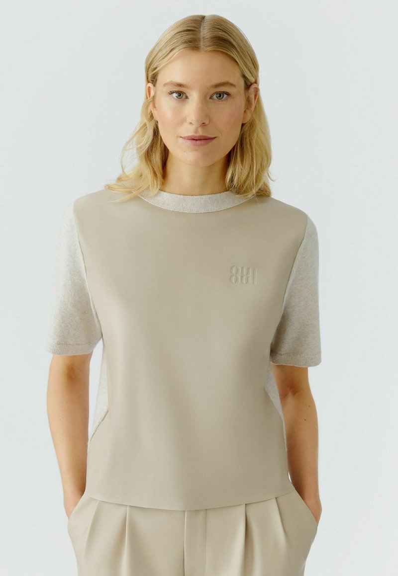 Beige bovenstuk met korte mouwen, met een gladde voorpaneel en gemarmerde mouwen zoals een trui. Subtiel logo detail op de borst.