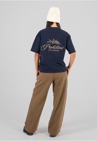 Camiseta de algodón azul marino de mangas cortas, con la inscripción dorada "Prohibido 10119 Berlín" en la parte posterior; combinada con pantalones de pierna ancha marrones.