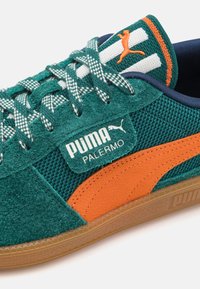 Tenisi verzi din suede cu accente portocalii, partea superioară din plasă și șireturi cu model. Dispune de un patch cu logo și o talpă din cauciuc texturată.