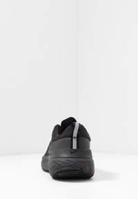 Zapatilla deportiva negra con parte superior de malla, suela de goma gruesa y detalles reflectantes en el talón y la entresuela. Diseño minimalista.