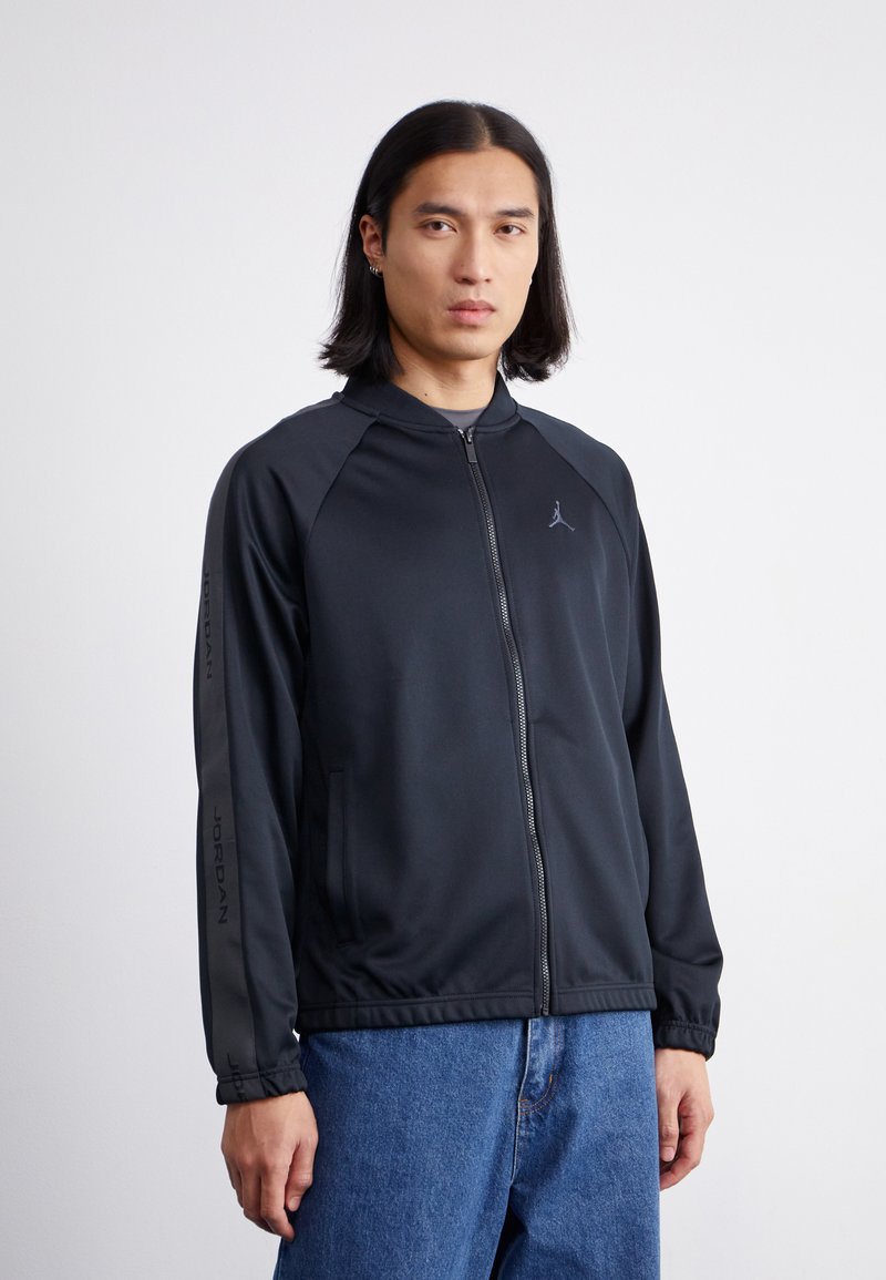 Sort zip-up sportjakke med raglanærmer, Jordan logo på brystet og ærmet, har sidelommer og en glat overflade. Denim jeans bæres nedenunder.