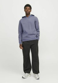 Lila Kapuzenpullover mit einer Fronttasche und Kordelzug, kombiniert mit schwarzen weit geschnittenen Hosen und silbernen Sportschuhen.