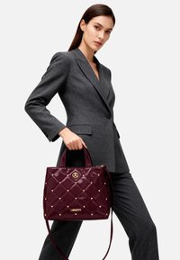 Borsa a tracolla in pelle trapuntata color bordeaux con dettagli in metallo dorato, dotata di chiusura con cerniera e doppi manici superiori. Decorata con borchie decorative.