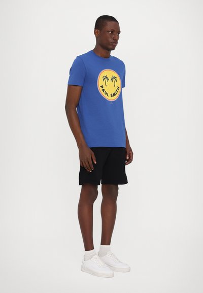 Zils kokvilnas T-krekls ar apļveida grafiku, kurā attēloti palmu koki un teksts "Paul Smith", kombinēts ar melniem šortiem un baltām sporta apaviem.
