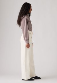 Γυναίκα με μακριά σγουρά μαλλιά στέκεται σε προφίλ φορώντας μια καρό μπλούζα με βολάν, λευκό παντελόνι φαρδύ με ψηλή μέση και μαύρα παπούτσια.