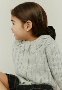 Vaaleanharmaa neulottu neuletakki, jossa on tekstuuriin perustuvat kukka-akcentit, napitus edessä ja ribbireunat. Musta ruudullinen hame näkyy alapuolella.