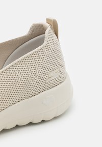 Chaussure slip-on en maille beige avec un intérieur doux, une semelle ergonomique et un extérieur texturé. Présente un logo discret sur le côté.