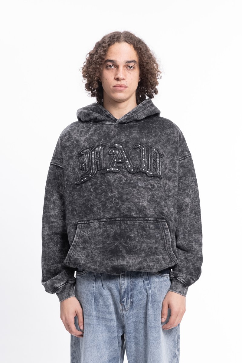 Jovem com cabelo cacheado, vestindo um hoodie cinza desbotado com a inscrição "MAD" e jeans leves de cintura baixa, em pé contra um fundo branco.