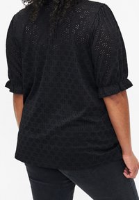 Blouse noire à manches courtes bouffantes avec un motif texturé de petits détails ajourés. Tissu doux et coupe décontractée, s'étendant jusqu'aux hanches.