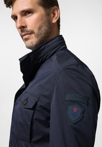 Granatowa kurtka outdoorowa z wysokim kołnierzem, kieszenią zapinaną na guziki oraz naszywkami na ramionach z wytłoczonymi logo i wzorem tarczy.