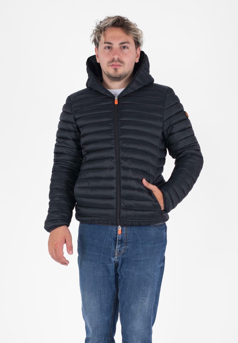 Zwarte pufferjack met horizontale stiksels, een ritssluiting aan de voorkant en een capuchon. Kenmerkt zich door oranje ritsaccenten en zijzakken. Draagt met blauwe jeans.